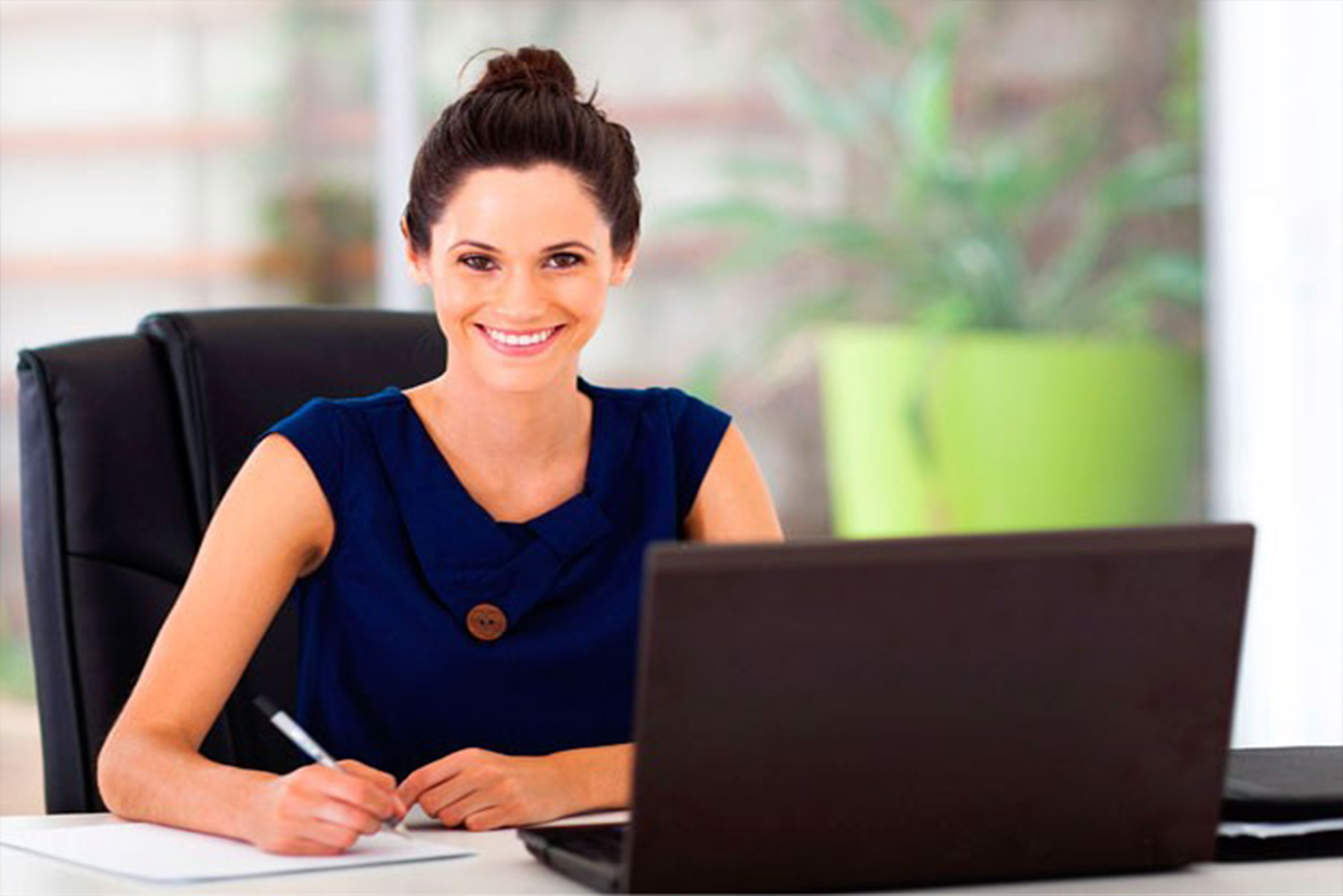 women sitting at table writing notes with open computer in front of her.